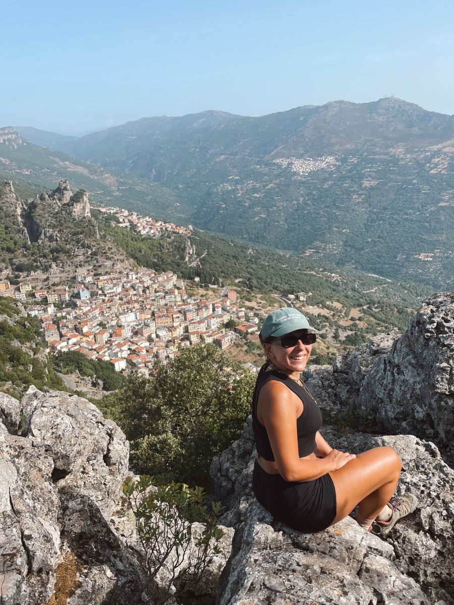 Una escursionista sorridente si siede su uno spuntone di roccia, ammirando lo spettacolare panorama del paese di Ulassai annidato tra i "tacchi" di calcare e la valle sottostante. Un'esperienza imperdibile per cosa vedere a Ulassai.
