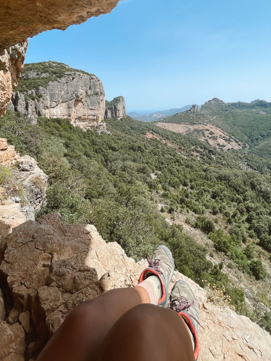 Vista in soggettiva dalle gambe e dalle scarpe da trekking di una escursionista seduta all'ingresso di una grotta naturale. Si affaccia su un paesaggio incontaminato di montagne calcaree e boschi mediterranei a Ulassai.